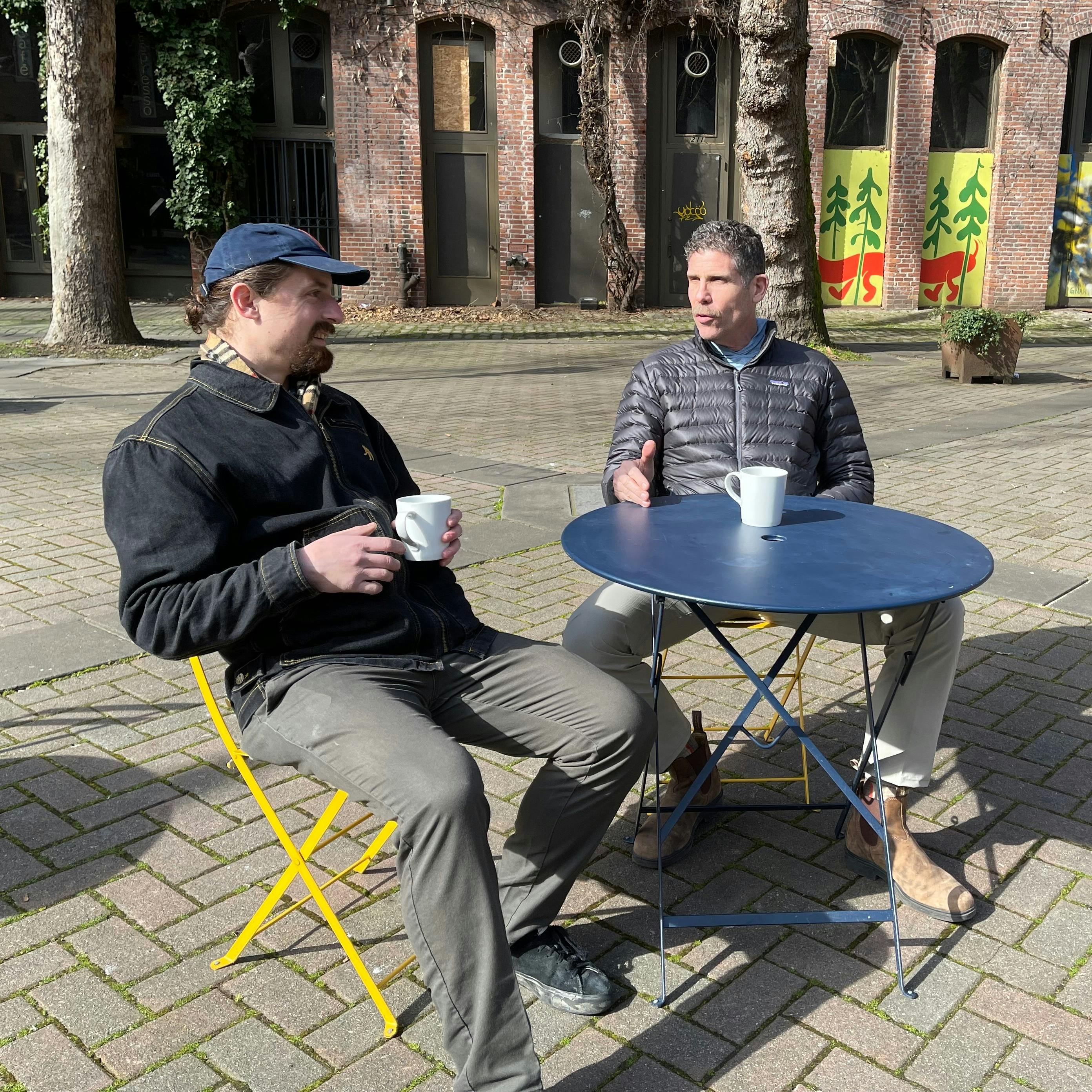 Dr. Shlomo Freiman and Tobias Coughlin-Bogue enjoying a coffee and a chat at a cafe table in a plaza on a sunny spring day